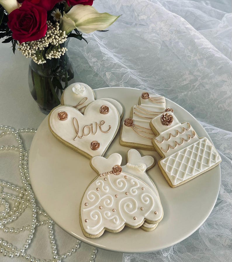 May include: Four decorated sugar cookies on a white plate. The cookies are decorated with white icing and gold accents. One cookie is shaped like a heart and says "love" in gold lettering. Another cookie is shaped like a wedding dress. The other two cookies are shaped like wedding cakes.