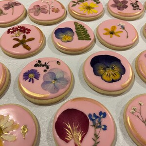 May include: A collection of pink sugar cookies decorated with pressed flowers and gold accents. The cookies feature a variety of flowers, including pansies, daisies, and ferns.