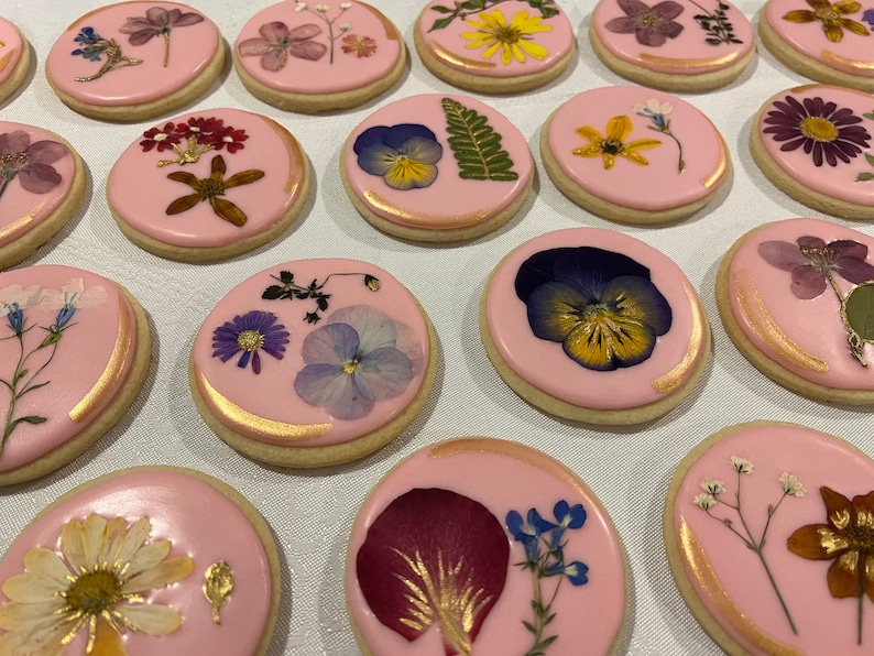 May include: A collection of pink sugar cookies decorated with pressed flowers and gold accents. The cookies feature a variety of flowers, including pansies, daisies, and ferns.