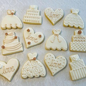 May include: A collection of 10 decorated sugar cookies featuring wedding themes. The cookies are decorated with white icing and gold accents. Some cookies are shaped like hearts and feature the word "love" in gold lettering. Other cookies are shaped like wedding cakes and wedding dresses.