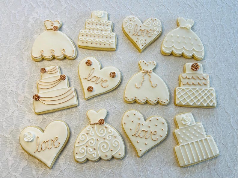 May include: A collection of 10 decorated sugar cookies featuring wedding themes. The cookies are decorated with white icing and gold accents. Some cookies are shaped like hearts and feature the word "love" in gold lettering. Other cookies are shaped like wedding cakes and wedding dresses.