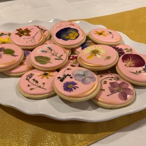 May include: A plate of pink sugar cookies decorated with pressed flowers. The cookies are oval shaped and have a gold shimmer.