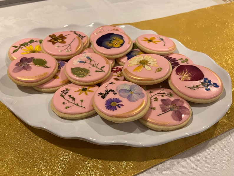 May include: A plate of pink sugar cookies decorated with pressed flowers. The cookies are oval shaped and have a gold shimmer.