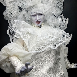 May include: A pale-skinned vampire doll with sharp fangs and dark red lips, wearing a white brocade dress, a large lace ruff collar, and an elaborate white tulle headpiece. The doll features pearl jewelry and long, puffed sleeves with lace trim.