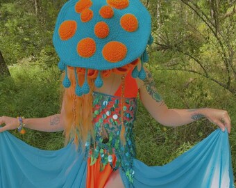 Könnte beinhalten: Ein skurriles Kostüm mit einem großen, türkisfarbenen gehäkelten Hut, der mit orangefarbenen Kreisen verziert ist. Das Outfit umfasst ein orangefarbenes und türkisfarbenes Kleid mit Pailletten und Stoffverzierungen sowie einen fließenden türkisfarbenen Rock. Das Ensemble ist vor dem Hintergrund üppigen Grüns inszeniert.
