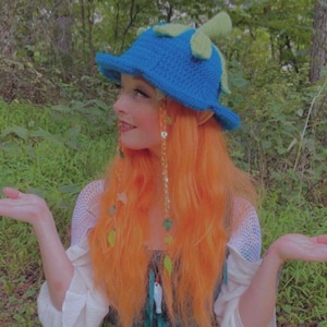 May include: A blue crochet hat with green leaf accents. The hat is worn by a person with orange hair and a white top. The background is a lush green forest. The hat is a whimsical accessory.