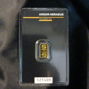 May include: A gold bar encased in a black plastic holder. The bar is labeled "1g Fine Gold 999.9" and "121409". The holder has the text "ARGOR-HERAEUS" and "The golden link".