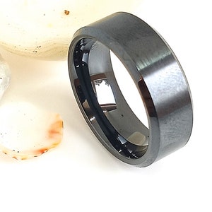 May include: A black ring with a flat top and a polished, reflective inner surface. The ring has a matte finish on the outer surface. The ring is set against a white background with a few seashells.