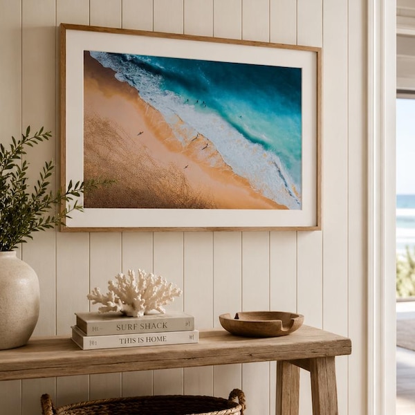 May include: A framed beach scene with turquoise water and golden sand hangs on a white paneled wall. Below, a wooden bench holds decorative items: a coral sculpture, books, and a wooden bowl. A woven basket and a jute rug complete the coastal decor.