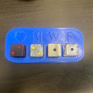 Pode incluir: Bandeja de dados de plástico azul com as letras M W F e um coração. Existem quatro dados na bandeja, um preto com dois pontos vermelhos, um branco com dois pontos amarelos, um cinza com um ponto preto e um prateado com um ponto preto.