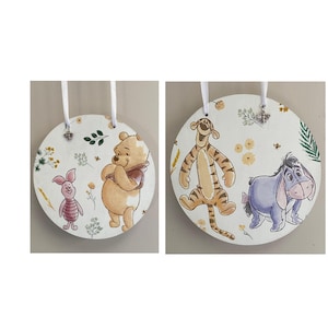 May include: Two circular wooden decorations featuring Winnie the Pooh characters. One shows Piglet and Winnie the Pooh, the other Tigger and Eeyore. Each has a white ribbon for hanging, with floral and bee accents.