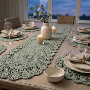 May include: A sage green crocheted table runner and matching round placemats with scalloped edges. The table setting includes white ceramic plates, beige cloth napkins with woven rings, clear glass wine glasses, a small bowl, and a vase.