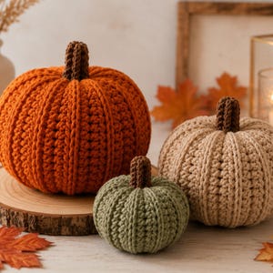 May include: Three crocheted pumpkins in orange, beige, and sage green, each featuring a textured ribbed pattern and a small brown stem. The pumpkins are arranged together, with the large orange one on the left, the beige one on the right, and the small green one in the front center.