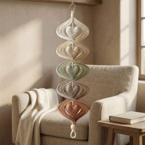 Puede incluir: Un móvil colgante vertical con cinco corazones de ganchillo en un degradado de colores desde crema hasta terracota. Cada corazón está encerrado en un marco de ganchillo calado en espiral, suspendido por un cordón central con un gancho.