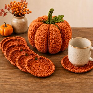 May include: A large orange crochet pumpkin with a green stem and leaf, accompanied by a stack of five round orange crochet pumpkin-shaped coasters and one additional coaster supporting a speckled ceramic mug on a wooden surface.