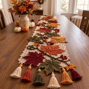 May include: A long, rectangular crochet table runner featuring an intricate cream-colored lace pattern adorned with three-dimensional crocheted autumn leaves in shades of red, orange, yellow, and green, finished with decorative tassels and wooden beads.