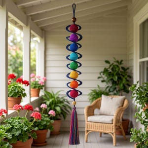 May include: A hanging mobile featuring a dark blue spiral structure adorned with nine crocheted spheres in a spectrum of colors including red, purple, blue, teal, green, yellow, and orange, finished with a long, multi-colored fiber tassel.