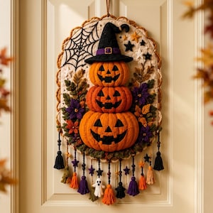May include: An oval quilted wall hanging featuring a stack of three orange jack-o'-lanterns wearing a black witch hat. The design includes a black spiderweb, purple and orange fabric flowers, and dangling beaded tassels with small ghost charms.