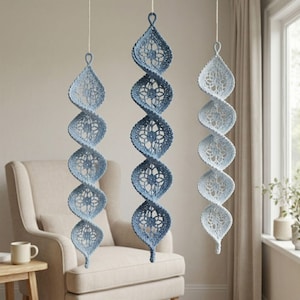 May include: Three hanging spiral ornaments crafted from crochet lace in varying shades of blue. Each piece features a repeating floral motif and a loop at the top for suspension. The ornaments are arranged vertically, displaying a gradient of blue tones.