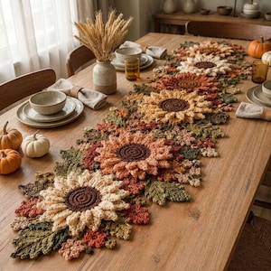 Op de afbeelding: Een lange, decoratieve gehaakte tafelloper met een herhalend patroon van grote zonnebloemen met donkerbruine harten en crème, perzik en roestkleurige bloemblaadjes, afgewisseld met gestructureerde groene, oranje en bruine gehaakte bladeren.