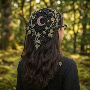 May include: A black crochet headscarf featuring an intricate vine and leaf pattern in olive green thread. The design includes a central crescent moon and small stars embroidered in a soft pink hue, with two dangling heart-shaped charms at the ends.