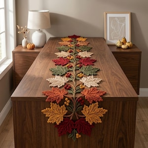May include: A crochet table runner featuring a central vine design adorned with textured maple leaves in shades of red, orange, yellow, green, and cream, accented with small clusters of round berries in red, yellow, and orange tones.