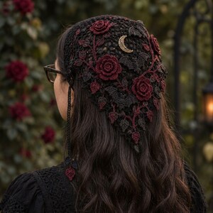 May include: A black triangular crochet headscarf featuring dark red rose appliqués, black crocheted bat motifs, and a gold-toned crescent moon charm. The headscarf has thin crocheted ties with small leaf-shaped accents at the ends.