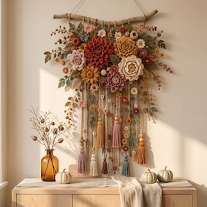 Può includere: Una decorazione da parete con una collezione di fiori all'uncinetto nei toni del rosso, arancione, giallo e crema, appesa a un ramo di legno naturale. La composizione include fili pendenti, perline di legno e nappe in tessuto colorate.