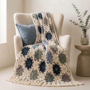 May include: A crochet blanket featuring a repeating floral motif in shades of cream, navy blue, sage green, and slate gray. The blanket is constructed from individual square panels joined together and finished with a fringe along the bottom edge.