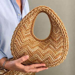 May include: A crescent-shaped handbag crafted from woven cord in a variegated tan and beige chevron pattern. The bag features a thick, braided border along the outer edge and a single integrated handle opening at the top, creating a structured silhouette.