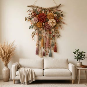 Può includere: Un grande arazzo da parete con una collezione di fiori all'uncinetto nei toni del rosso, arancione, giallo e crema, fissato a un ramo di legno naturale. Lunghe corde in macramè con perline di legno e nappe colorate pendono sotto.