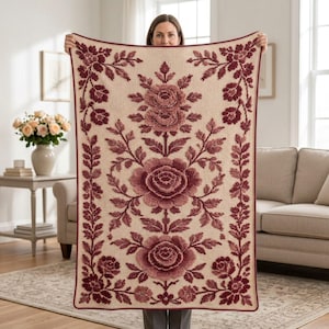 May include: A rectangular throw blanket featuring a cream-colored base with a textured, raised floral pattern in shades of deep burgundy and maroon. The design includes three large central rose motifs and intricate vine borders along the edges.