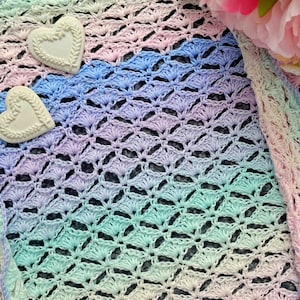 May include: A crocheted shawl in a rainbow of pastel colors, including pink, blue, and green. The shawl features a delicate shell stitch pattern and is adorned with two heart-shaped embellishments. A pink flower is in the background.