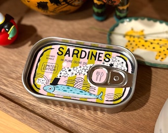 Puede incluir: Lata rectangular de sardinas con una etiqueta colorida. La etiqueta muestra la palabra "Sardines", una ilustración de pez azul y "In Pure Olive Oil". La lata es una edición limitada de pegatinas. Peso neto 105 gramos.