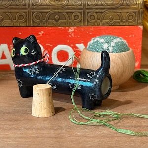 Puede incluir: Un sujetador de agujas con forma de gato negro de cerámica con detalles de estrellas blancas y ojos verdes. Un corcho y un cojín de alfileres de cactus están cerca. El hilo verde está enhebrado a través de la aguja, lo que sugiere un tema de costura o bordado.