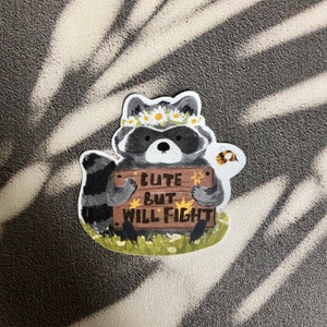 Puede incluir: Un adorable mapache con una corona de flores sostiene un cartel que dice "Cute but will fight". Una peque&ntilde;a abeja vuela junto al mapache.