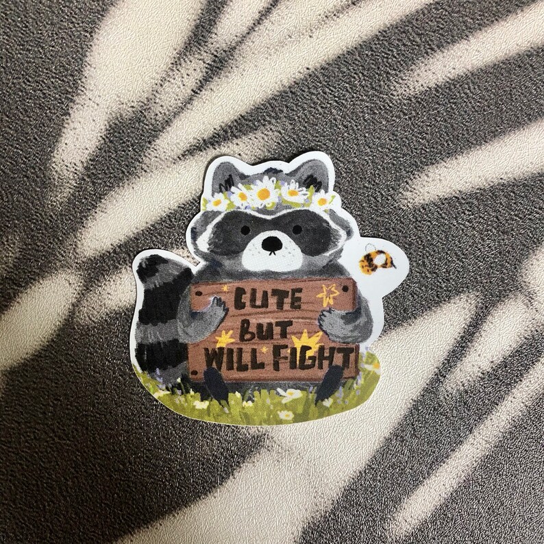 Puede incluir: Un adorable mapache con una corona de flores sostiene un cartel que dice "Cute but will fight". Una peque&ntilde;a abeja vuela junto al mapache.