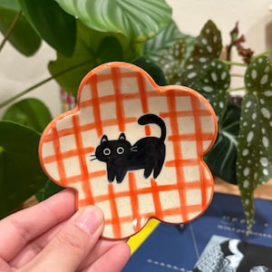 Könnte beinhalten: Eine blütenförmige Keramikschale mit einem orange-weiß karierten Muster. In der Mitte ist eine schwarze Katze aufgemalt. Die Schale wird in der Hand einer Person gehalten, mit grünen Pflanzen im Hintergrund.
