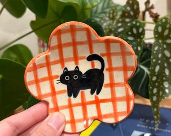 May include: A flower-shaped ceramic dish with an orange and white checkered pattern. A black cat is painted in the center. The dish is held in a person's hand, with green plants in the background.