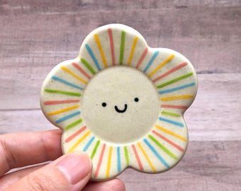 May include: A ceramic flower-shaped trinket dish with a smiling face in the center. The dish is off-white with colorful stripes radiating from the center. The stripes include pink, yellow, blue, and green.