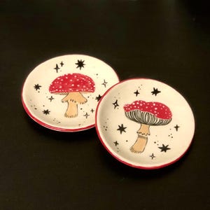 May include: Two small white ceramic dishes with red rims. Each dish features a black and white illustration of a red and white mushroom with white stars on a black background.