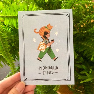 May include: A blue illustrated card with a woman walking with a cat on her head. The woman is holding a cup of coffee and a tote bag. The text on the card reads "I'm controlled by cats."