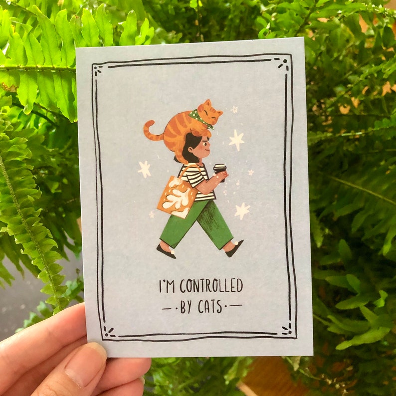 May include: A blue illustrated card with a woman walking with a cat on her head. The woman is holding a cup of coffee and a tote bag. The text on the card reads "I'm controlled by cats."