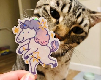 May include: A sparkly iridescent unicorn sticker with a pink mane and tail, held by a hand in front of a tabby cat.