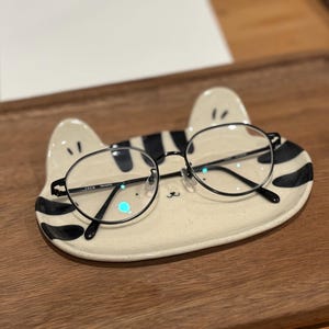 以下が含まれることがあります： 丸い黒縁メガネが、猫の形をした陶器の皿の上に置かれています。 皿はオフホワイトで、黒い縞模様と猫の耳が付いています。 メガネは細い黒いフレームと透明なレンズです。 皿は木製の表面に置かれています。