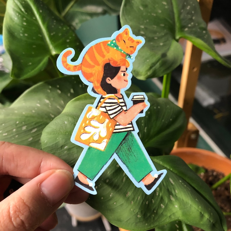 May include: A sticker featuring a cartoon illustration of a person with a cat on their head, carrying a tote bag and a cup of coffee. The person is wearing a striped shirt and green pants. The cat is orange and white.