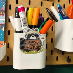 Puede incluir: Porta l&aacute;pices de cer&aacute;mica blanca con una pegatina de mapache que dice "Cute but will fight". El porta l&aacute;pices est&aacute; lleno de marcadores y bol&iacute;grafos de varios colores.