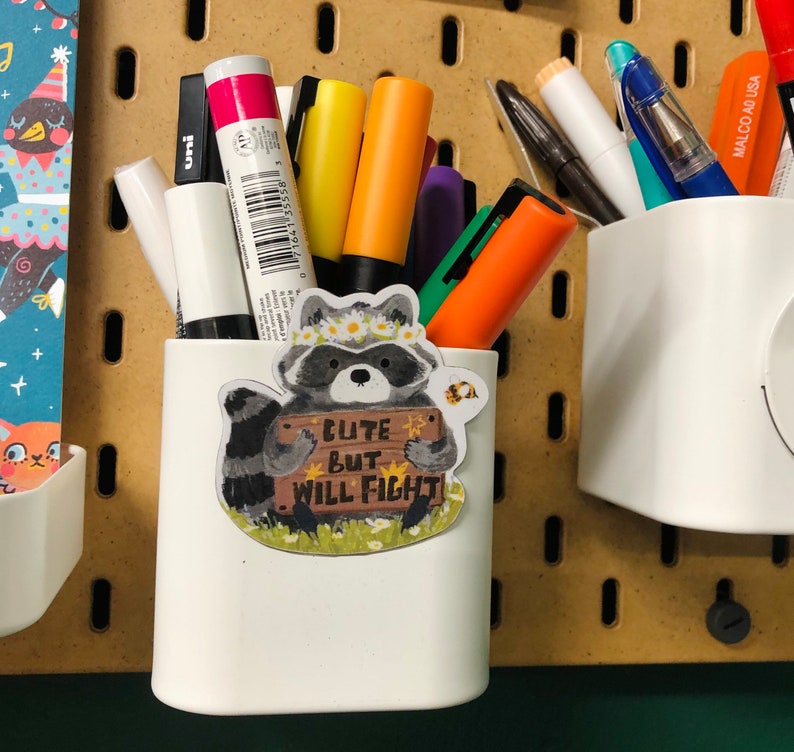 Puede incluir: Porta l&aacute;pices de cer&aacute;mica blanca con una pegatina de mapache que dice "Cute but will fight". El porta l&aacute;pices est&aacute; lleno de marcadores y bol&iacute;grafos de varios colores.