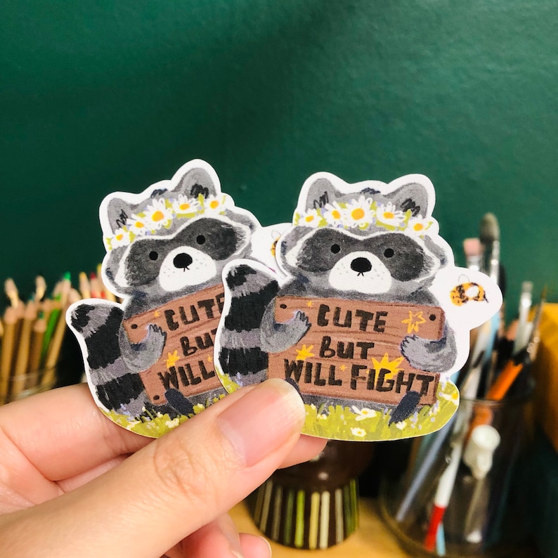 Puede incluir: Dos pegatinas ilustradas de mapaches con diademas florales y un cartel de madera que dice "Cute but will fight".