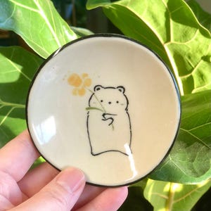 May include: A small white ceramic dish with a black rim features a hand-drawn illustration of a bear holding a yellow flower.